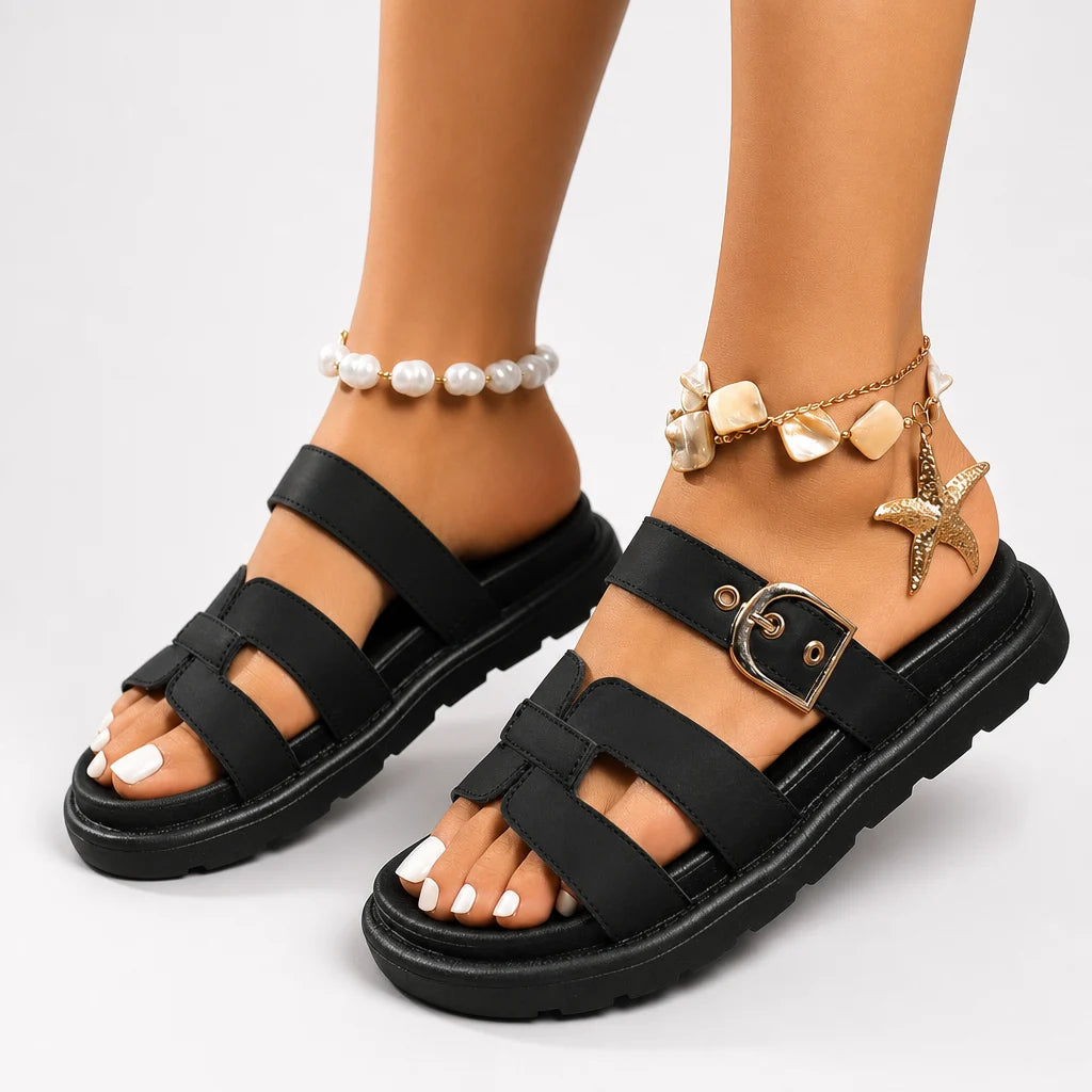 Black strappy platform sandals with buckle detail worn on feet in a white studio.