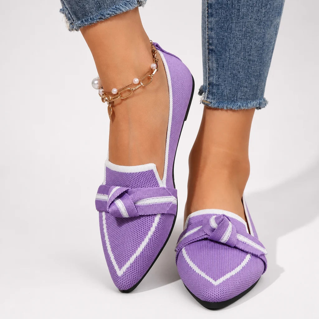 Purple pointed flats with bow details worn by a model in a bright studio.
