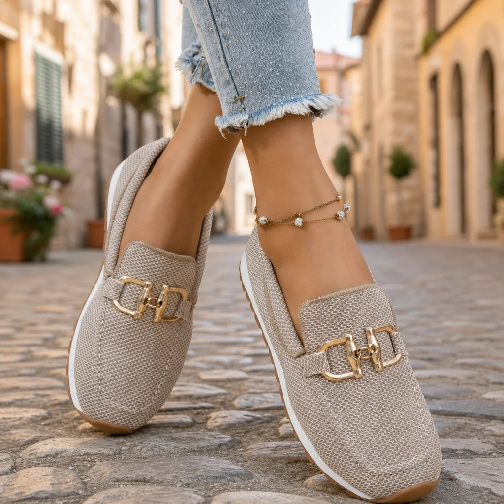 Beige woven loafers with gold buckles worn on feet in a sunny Italian street.