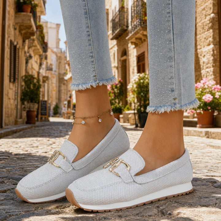 Light gray textured loafers with gold buckles worn on a sunny Italian street.