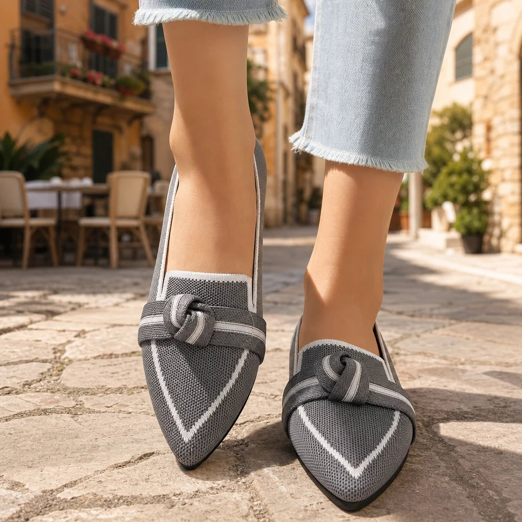 Gray knit pointed loafers with white trim and bow straps worn on a sunny Italian street.