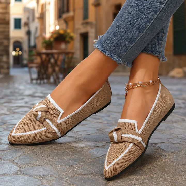 Beige pointed loafers with white trim and bow worn on feet in a sunny Italian street.