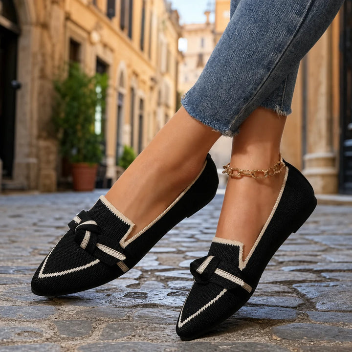 Black knit pointed loafers with cream trim and bow accents worn with cropped jeans.